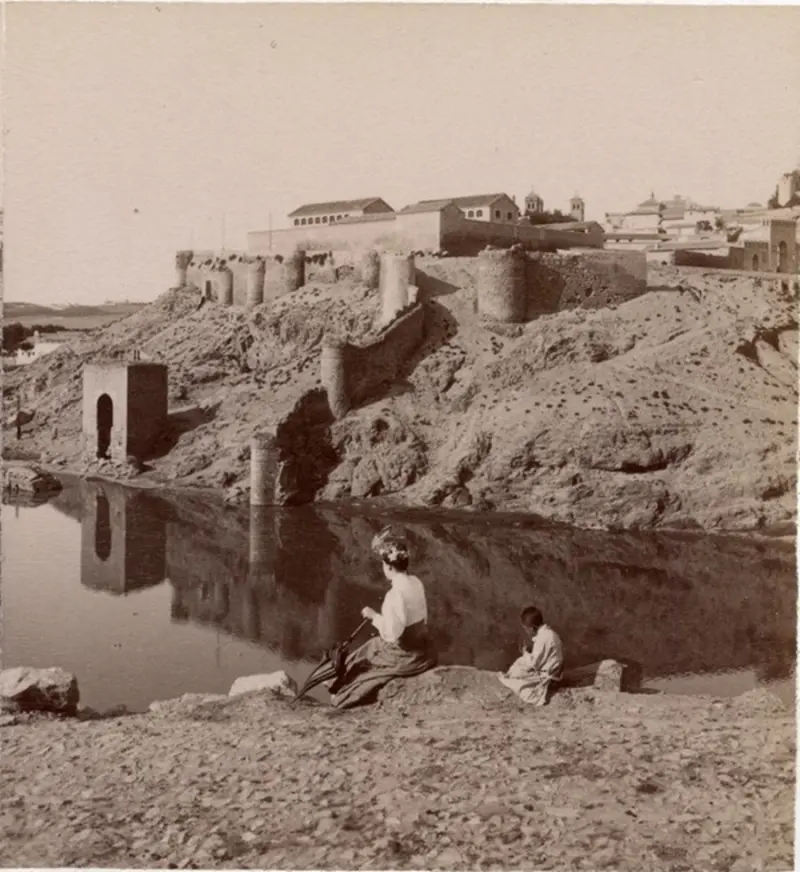 Mujer con sombrilla y un niño con el Baño de la Cava al fondo. 