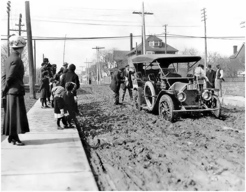 Crowd on sidewalk watching a automobile mired in the mud