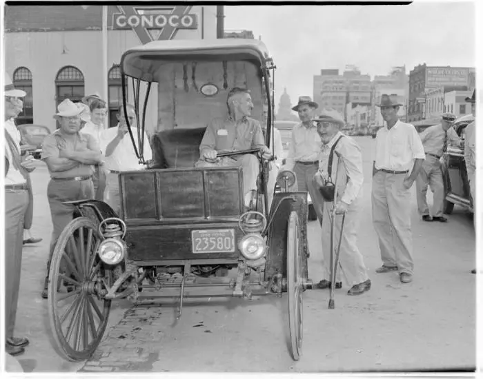 Man in old car, surrounded by small crowd. 