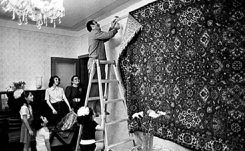 The head of the family, under the enthusiastic glances of his relatives, nails a luxurious carpet to the wall